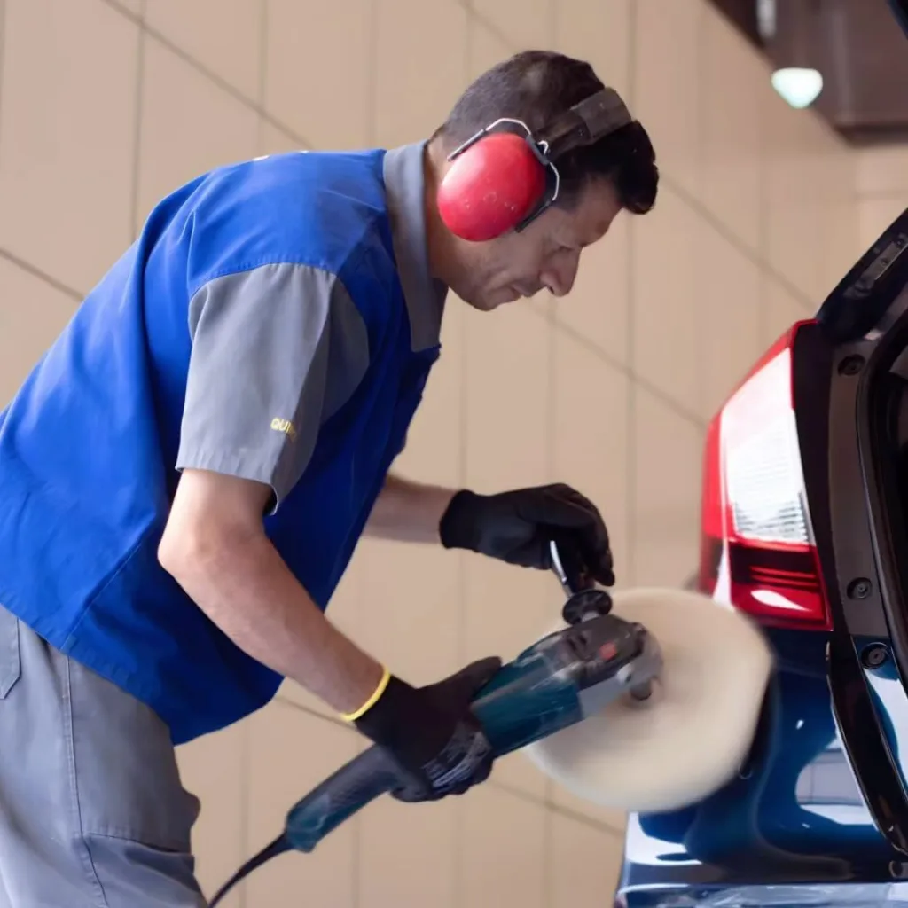 Profissional de perfil usando protetor auricular vermelho, luvas pretas e uniforme azul com cinza enquanto opera uma politriz com boina de lã branca na lateral de um carro azul escuro.